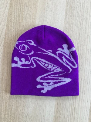 Lila mössa med grodmotiv - Säljer en lila mössa med grodmotiv på framsidan. Ny. Unisexmodell. Har fler färger och skickar gärna med samfrakt. One size 