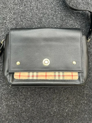 Svart axelväska från Burberry - Snygg svart axelväska i läder från Burberry med klassiskt rutigt mönster på framsidan. Väskan har guldfärgade detaljer och justerbar axelrem. Perfekt för dig som gillar stilrena accessoarer med en twist.