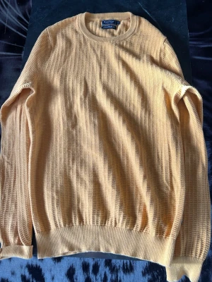 Gul stickad tröja från East West - Säljer en gul stickad tröja från East West Athletic Supply Co. Tröjan har rund halsringning, ribbade muddar och nederkant samt ett diskret rutmönster i stickningen. Perfekt för dig som gillar en enkel men snygg stil.