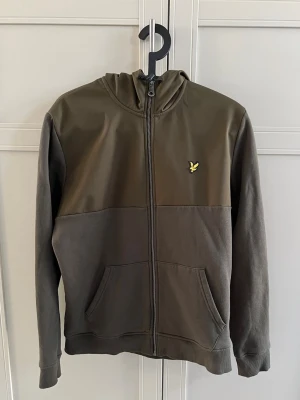 Olivgrön hoodie med dragkedja - Snygg olivgrön hoodie från Lyle & Scott med dragkedja framtill och klassisk gul logga på bröstet. Hoodien har delad känguruficka, ribbade muddar och en tvådelad design med olika nyanser av grönt. Perfekt för en avslappnad stil.