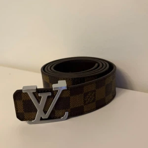 Brunt bälte - Säljer ett brunt rutigt bälte med den klassiska loggan i silver på spännet. Bältet är gjort i skinn och har ett stilrent, lyxigt utseende med mörkbruna och ljusbruna rutor. Perfekt accessoar för att lyfta din outfit.