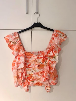 Blommig volangtopp med breda axelband - Köpte denna av en annan men den passade tyvärr inte mig!🌸 Supersöt topp med blommigt mönster i rosa, rött och vitt. Modellen har breda axelband och volangdetaljer längs axlar och sidor. Smockad kropp för en tight passform och luftig känsla, perfekt för varma dagar.