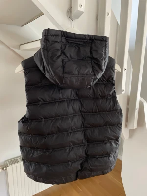 Svart dunväst med huva - Snygg svart dunväst med huva och dragkedja framtill. Västen har två fickor med dragkedja och quiltad design. Perfekt för lager-på-lager och ger en sportig look. Materialet är glansigt och puffigt för extra värme.