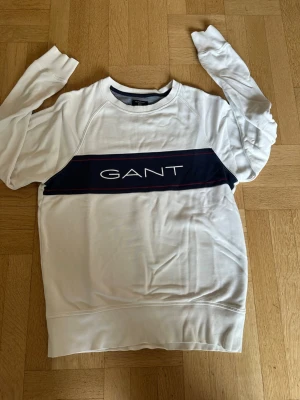 Vit sweatshirt från GANT - Snygg vit sweatshirt från GANT med marinblå panel och stor logga över bröstet. Tröjan har långa ärmar, ribbade muddar och rund halsringning. Perfekt för en avslappnad och stilren look.