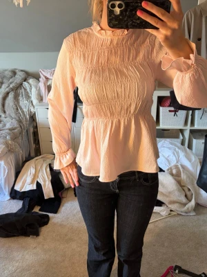 Ljusrosa blus från Vero Moda - Supersöt ljusrosa blus från Vero Moda med hög krage och volangdetaljer. Blusen har långärmad design, smockad struktur och lätt utsvängd midja som ger en feminin siluett. Tillverkad i ett tunt, strukturerat tyg som känns luftigt och bekvämt.