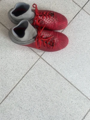 Röda fotbollsskor med högt skaft - Säljer ett par röda fotbollsskor med högt, stickat grått skaft och röda snören. Skorna har ett mönster i vitt på ovansidan och är designade för bra grepp på planen. Perfekta för dig som vill ha stil och funktion på matchen.
