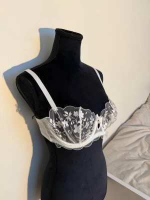 Vit spets-bh med blommönster - Söt vit bh med transparent spets och broderade blommor framtill. Bh:n har justerbara axelband och en liten rosett i mitten. Kuporna är lätt vadderade och ger en feminin look. Perfekt för dig som gillar romantiska detaljer och elegant design.