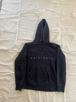 Svart hoodie med tryck från Originals - Snygg svart hoodie från Originals med vit text på bröstet. Klassisk känguruficka framtill och justerbar huva med snören. Tillverkad i mjukt material som känns skönt mot huden. Perfekt för en avslappnad och trendig stil.