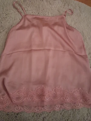 Rosa nattlinne med spetsdetaljer - Sött nattlinne i ljusrosa med tunna axelband och vacker spetskant längst ner. Tillverkat i ett mjukt, glansigt material som känns skönt mot huden. Perfekt för mysiga kvällar hemma.