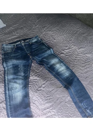 Blå slitna  jeans med detaljer - Säljer ett par blå jeans med slitningar och vita färgstänk för en cool, trendig look. Jeansen har klassisk femficksdesign och kontrastsömmar. Perfekta för dig som gillar streetstyle och vill sticka ut lite extra.