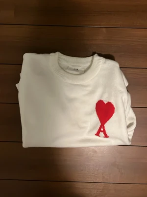 Vit stickad tröja från Ami Paris - Säljer en vit stickad tröja från Ami Paris med röd hjärtlogga och bokstaven A på bröstet. Tröjan har rund halsringning och ribbade muddar. Perfekt för dig som gillar stilrena plagg med en twist.