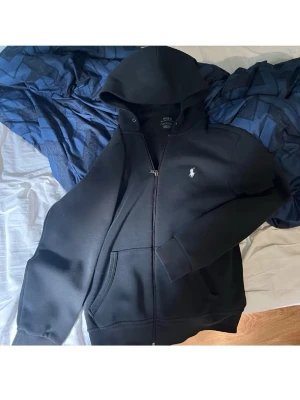 Svart hoodie från Polo Ralph Lauren - Säljer en svart hoodie från Polo Ralph Lauren med dragkedja och klassisk vit logga på bröstet. Hoodien har huva, fickor framtill och ribbade muddar. Perfekt för en chill och stilren look. Materialet är mjukt och skönt, troligtvis bomullsblandning.