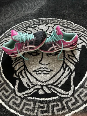 Asics sneakers svart rosa turkos - Snygga Asics sneakers i svart mesh med rosa och turkosa detaljer. Skorna har vita loggor på sidorna, turkosa skosnören och en bekväm sula med rosa och vita inslag. Perfekta för dig som gillar färg och sportig stil.