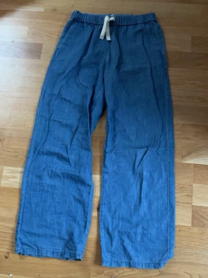 Blå linnebyxor med dragsko - Säljer ett par blå linnebyxor med avslappnad passform och breda ben. Byxorna har resår i midjan och vit dragsko för enkel justering. Perfekta för en chill och bekväm stil. Materialet är lätt och luftigt linne.