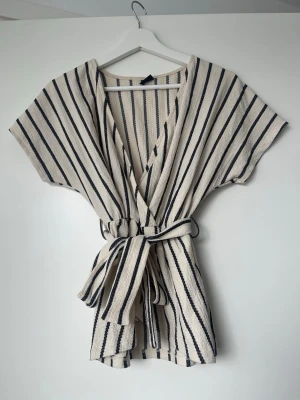 Randig omlottopp med knytband - Säljer en beige och mörkblå randig omlottopp med knytband i midjan. Toppen har korta ärmar och ett avslappnat, loose fit. Perfekt för en chill och stilren look.