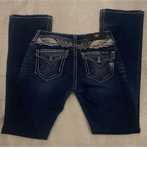 Mörkblå Miss Me jeans med brodyr - Säljer ett par mörkblå jeans från Miss Me med snygga kontrastsömmar och broderade detaljer på bakfickorna. Jeansen har raka ben, låg midja och klassisk femficksdesign. Materialet är stretchigt denim för skön passform. Byxorna saknar en knapp på fikan på baksidan men inget som man tänker på och dom är även lite slitna ner till som beror på att dom va lite för långa för mig annars sitter dom jättefint. Köpte dom här på Plick för 1100 då d syndes att dom va använda.