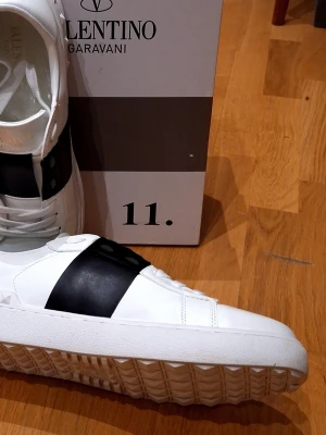 Valentino Open - Snygga vita sneakers från Valentino Garavani med en bred svart rem över mitten. Skorna har en chunky sula med mönster och detaljerade nitar längs kanten. Tillverkade i läder för en lyxig känsla och stilren look.