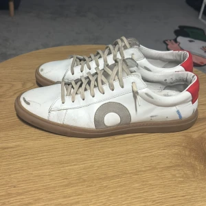 Vita sneakers med röd häl och grå detalj - Snygga vita sneakers med röd häl och en stor grå cirkeldetalj på sidan. Skorna har en klassisk låg profil med gummisula. Materialet är läder och designen sleten och snygg 