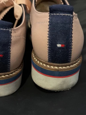 Rosa loafers från Tommy Hilfiger - Snygga rosa loafers i läder från Tommy Hilfiger med breda satinband som snörning, vit sula med blå och röd rand samt dekorativt band med logga baktill. Perfekta för dig som vill ha en preppy och trendig look.