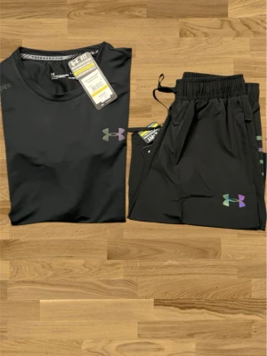  träningsset Under Armour - Snyggt svart träningsset från Under Armour med t-shirt och shorts. Båda plaggen har logga i regnbågsfärger och är tillverkade i ett lätt, snabbtorkande funktionsmaterial. Perfekt för gymmet eller löpning. T-shirten har rund hals och kort ärm, shortsen har resår i midjan.