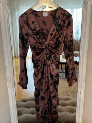 Långklänning i Silke med blommönster - Snygg långklänning i glansigt i Silke från Noa Noa med svart blommönster på puderrosa botten. Klänningen har lång ärm, v-ringning och en twistad detalj framtill som ger en cool draperingseffekt. Perfekt för dig som vill sticka ut med en elegant och trendig look. Köpt för 3300kr