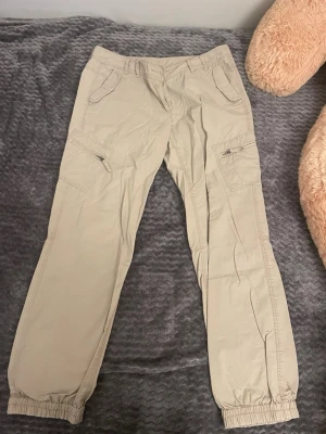 Beige cargobyxor från Savage - Snygga beige cargobyxor från Savage med avslappnad passform och resår vid benslut. Byxorna har flera praktiska fickor, inklusive dragkedjeförsedda sidofickor. Tillverkade i ett lätt och bekvämt material, perfekta för en casual streetwear-look.