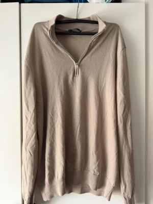 Beige halvzip tröja - Säljer en beige långärmad tröja med halvzip och hög krage. Tröjan har en avslappnad passform och ribbade muddar vid ärmslut och nederkant. Perfekt för lager på lager och enkel att matcha med olika outfits.