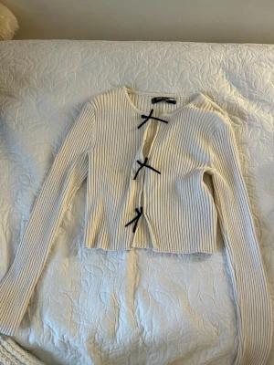 Vit ribbad kofta med svarta rosetter - Trendig vit ribbad kofta med långa ärmar och tre svarta rosetter som knäppning framtill. Kortare modell med rund halsringning, perfekt för att lyfta en outfit. Materialet är mjukt och stretchigt för en bekväm passform.