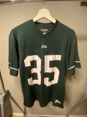 Grön sporttröja med nummer 35 - Mörkgrön sporttröja från New Garments med vitt tryck av siffran 35 på framsidan och texten SEATTLE på ryggen. Tröjan har V-ringning, korta ärmar och vita detaljer på ärmarna. Perfekt för dig som gillar amerikansk sportstil.