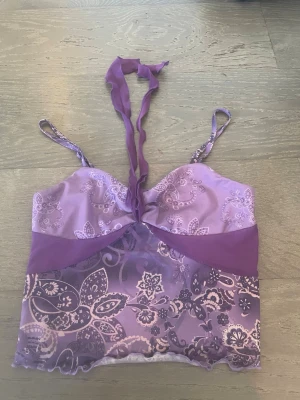 Lila mesh toppmed spetsdetaljer - Säljer en lila topp i mesh med tunna axelband och knytband framtill. Plagget har ett blommigt spetsmönster och transparent nederdel. Materialet är lätt och stretchigt, perfekt för dig som gillar feminina detaljer och en unik look. Kan användas på olika sätt. Köpt sacond hand