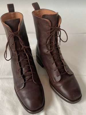 Bruna snörade läderstövletter - Snygga bruna stövletter i äkta läder med snörning framtill och fyrkantig tå. Skorna har en klassisk siluett med låg klack och dragflikar bak för enkel påtagning. Perfekta för dig som gillar tidlös och stilren design.