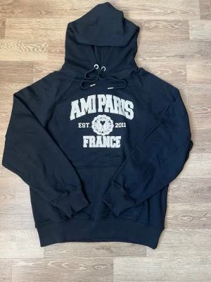 Svart hoodie från Ami Paris - Snygg svart hoodie från Ami Paris med stor vit text på bröstet och tryck med 'AMI PARIS FRANCE' samt 'EST. 2011'. Klassisk huva med dragsko och ribbade muddar. Perfekt för en avslappnad och trendig look.