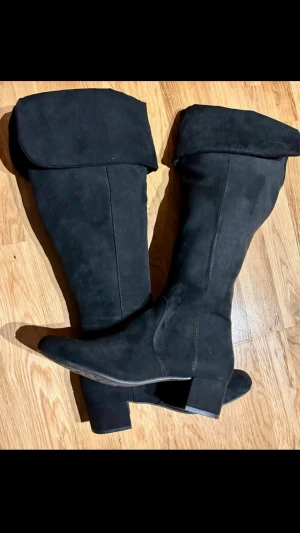 Svarta knähöga mockastövlar - Snygga svarta knähöga stövlar i mocka med bred klack och dragkedja på insidan. Stövlarna har en vikt kant upptill som ger en cool och trendig look. Perfekta till höst- och vinteroutfits.