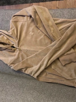 Beige zip hoodie från Juicy Couture  - Säljer en beige hoodie från Juicy Couture med dragkedja framtill och två stora fickor. Tillverkad i mjukt material med broderad logga på bröstet. Perfekt för chill dagar och enkel att matcha med jeans eller mjukisbyxor.