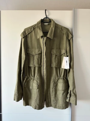 Grön overshirt från Gina Tricot - Stilren grön overshirt från Gina Tricot med fyra stora fickor framtill, axelklaffar och klassisk krage. Jackan har knappar vid ärmsluten och dragsko i midjan för en snygg passform. Perfekt lager på lager-plagg för vår och höst.