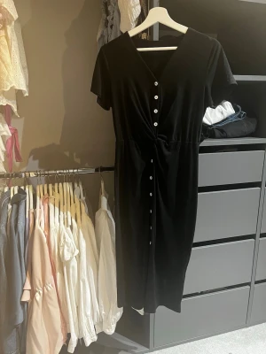 Svart långklänning med knappar - Stilren svart långklänning med v-ringning och korta ärmar. Klänningen har dekorativa vita knappar framtill och en smickrande midjeskärning. Perfekt för dig som gillar enkel men elegant stil.