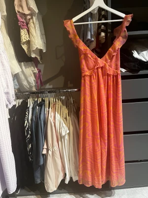 Rosa-orange långklänning med volang - Säljer en färgstark långklänning i rosa och orange med volangdetaljer på axelbanden och snygg knytning i ryggen. Klänningen har ett unikt mönster och är perfekt för sommardagar. Luftigt material och v-ringad framtill.