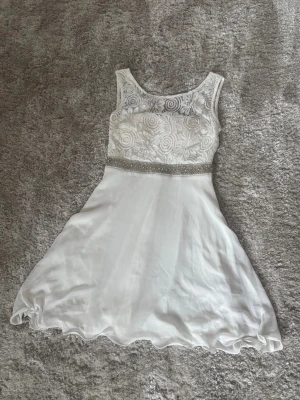 Vit kort klänning med spetsdetaljer - Söt vit kortklänning med broderade spetsdetaljer upptill och markerad midja med glittrigt band. Kjolen är lätt utsvängd och har vågig kant. Klänningen är ärmlös och har dragkedja i ryggen. 