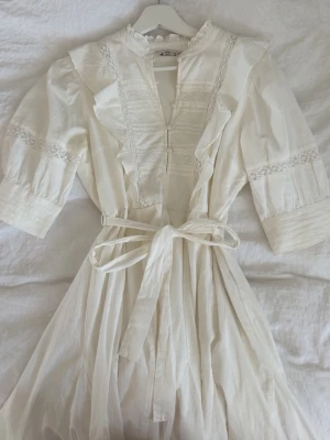 Vit omlottklänning med spetsdetaljer - Superfin vit klänning från ONLY med vackra spetsdetaljer, volanger och knytband i midjan. Klänningen har trekvartsärm, v-ringning och knappar framtill. Perfekt för sommarens alla tillfällen.