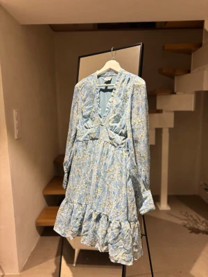 Blommig klänning från Aware - Blommig klänning från Aware by Vero Moda med blommigt mönster i blått och grönt. Klänningen har volangkant nertill, v-ringning och långa ärmar. Perfekt för vår och sommar, tillverkad i mjuk polyester. Se även på bilden att det är lite trasig vid vringningen! 🌸