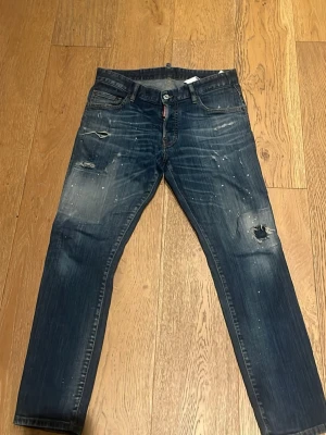 Blå slitna jeans från Dsquared2 - Säljer ett par blå jeans från Dsquared2 med distressed detaljer, hål på knät och coola färgstänk. Jeansen har klassisk femficksmodell, normal midja och raka ben. Perfekta för en avslappnad streetwear-look.