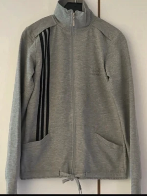 Grå zip-hoodie från Adidas - Snygg grå zip-hoodie från Adidas med svarta ränder på ena sidan och stort Adidas-tryck på ryggen. Tröjan har hög krage, dragkedja hela vägen, två fickor och snörning nedtill. Perfekt för en sportig look och chill dagar.