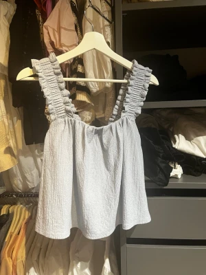 Ljusblå linnetopp med volangband - Supersöt ljusblå linnetopp med breda volangprydda axelband och knyt i ryggen. Toppen har en luftig och avslappnad passform, perfekt för varma dagar. Materialet känns lätt och svalt mot huden.