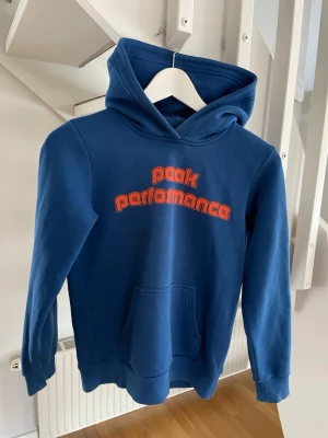 Blå Peak Performance hoodie - Snygg blå hoodie från Peak Performance med röd-orange logga på bröstet. Klassisk känguruficka framtill och stor huva. Tillverkad i mjukt material som känns skönt mot huden. Perfekt för en avslappnad och sportig stil.