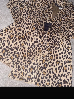 Leopardmönstrad kofta med knytband - Snygg leopardmönstrad kofta i beige och bruna toner med svart knytband i midjan. Koftan har lång ärm och är stickad, perfekt för dig som vill sticka ut lite extra. Passar till många olika stilar och är superenkel att matcha. Peplum passform 