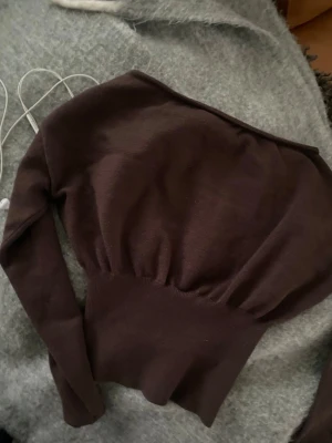 Brun off shoulder stickad topp - Säljer en brun stickad topp med off shoulder-design och lång ärm. Tröjan har en bred, ribbad midja som ger en snygg siluett och ballongliknande överdel. Perfekt för dig som gillar trendiga och bekväma plagg i mjukt material.