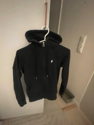 Svart hoodie från Peak Performance - Säljer en svart hoodie från Peak Performance med dragkedja framtill och liten logga på bröstet. Hoodien har justerbar huva med snören och är tillverkad i mjukt material som passar perfekt till chill eller streetwear. Klassisk och stilren design. Betalning via vanlig swish 
