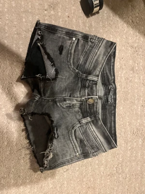 Grå slitna jeansshorts - Säljer ett par grå denimshorts med slitna detaljer och råa kanter. Shortsen har fem fickor, knappgylf och en cool, tvättad look. Perfekta för dig som gillar en edgy stil.