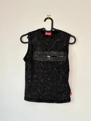 Svart ärmlös topp från Diesel - Cool svart ärmlös topp från Diesel med tvättad look och en stor bröstficka med dragkedja. Toppen har en edgy vibe och är perfekt för dig som gillar streetstyle. Materialet känns som bomull och toppen har en normal passform. Nypris 2000