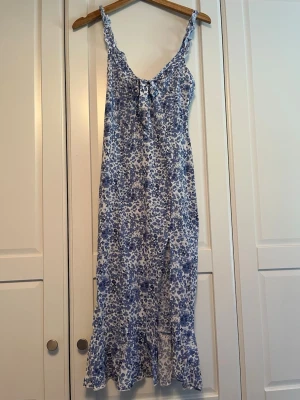 Blåvit blommig långklänning - Söt långklänning med blått och vitt blommönster. Klänningen har smala axelband med volangdetaljer och en dekorativ knytning framtill. Tillverkad i ett lätt och luftigt material, perfekt för varma dagar.
